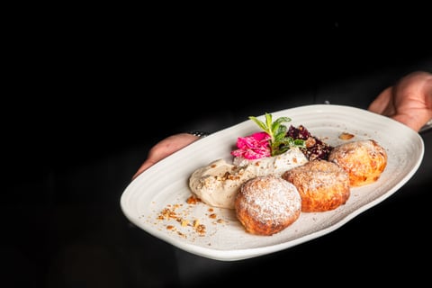 A waiter presents a plated dessert of three golden, powdered-sugar-dusted curd dumplings (Túrógombóc) served with cream, jam, and mint garnish on a white platter against a black background