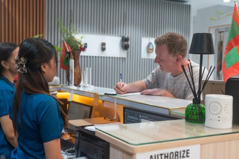Man in gray shirt at reception desk signing documents while two women in blue shirts wait in front of counter in office.