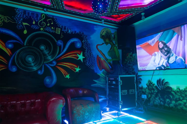 Neon-lit karaoke or entertainment room with colorful wall murals, speakers, screens, and LED lighting in blue, purple, and red tones