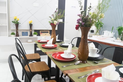 A bright dining room with a long table set with red placemats, white plates, and a green runner. Wooden chairs line the table, which is decorated with potted flowers and wooden vases containing yellow and pink blooms.