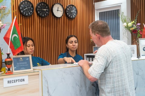Man at hotel reception desk speaking with two female staff members. Multiple clocks on wooden wall above, flag visible in background.