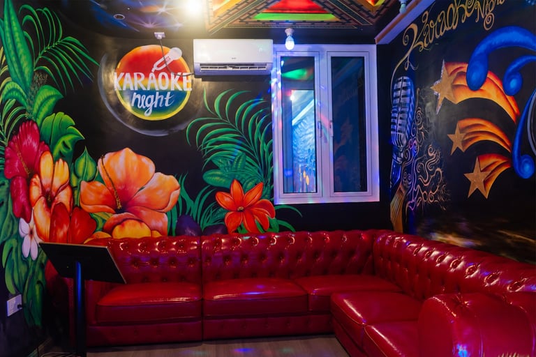 Vibrant karaoke lounge with neon signage, red leather seating, and colorful floral and abstract wall art illuminated by blue and yellow lights