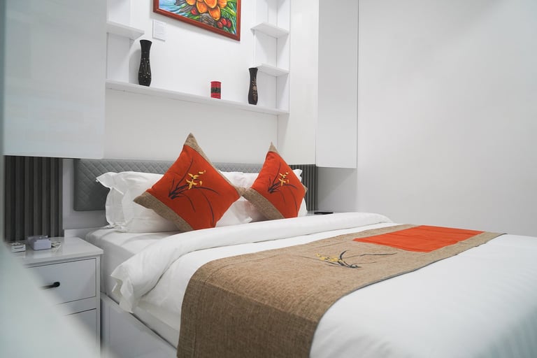 Modern bedroom with white bed, red decorative pillows, brown runner, and minimalist white shelving with decor above headboard